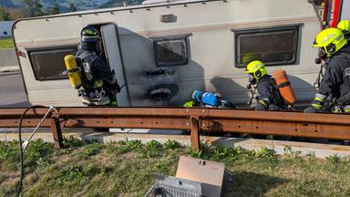 Roulotte in fiamme sull'autostrada del Brennero con vigili del fuoco al lavoro, fumo denso e traffico bloccato, immagine di approfondimento